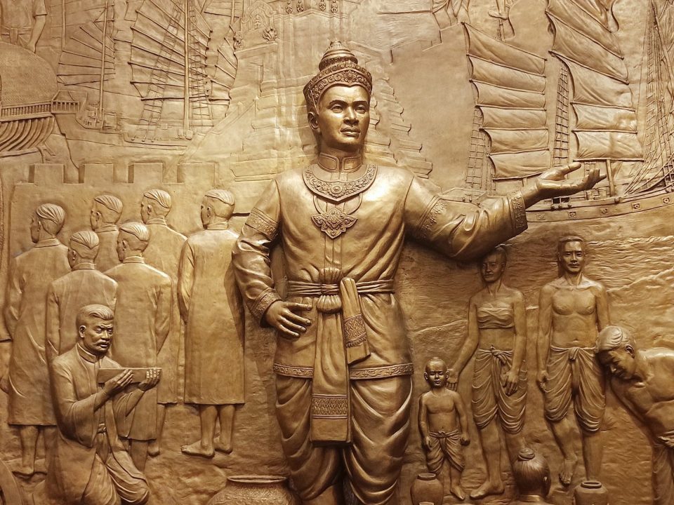 Gilded bas-relief of a crowned king with outstretched arm, surrounded by courtiers, villagers and sailing junks.