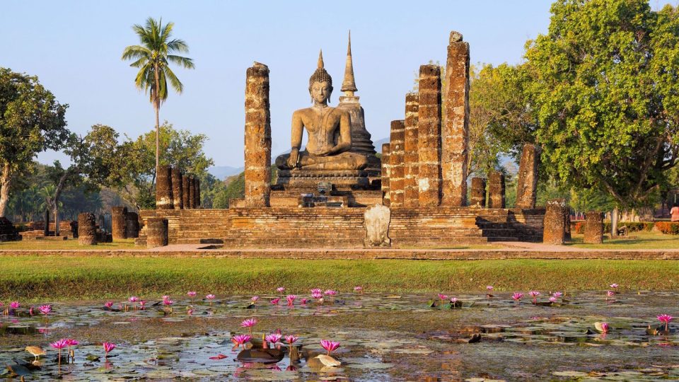 Seated brick and stucco Buddha between ruined columns, before a chedi, with a pink water-lily pond in the foreground.