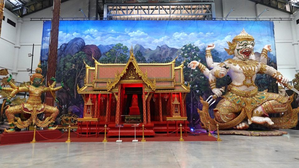 Décor de khon au centre d'apprentissage de Bang Pa-in : un pavillon sala doré encadré par deux géants du Ramakien, dont l'un, à la peau blanche et aux bras multiples, brandit des armes. — © Eric Monteil Décor de scène dans un hangar : pavillon thaï doré et rouge devant une toile de montagnes, entre deux grandes statues de géants.