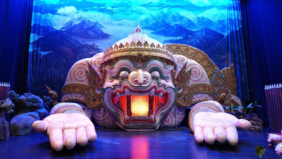 Theatre stage featuring a giant crowned demon head with open hands and a lit mouth, before a blue mountain backdrop.