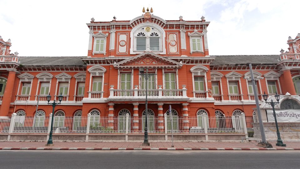 Façade of a European-style orange-red building with white stucco trim, green shutters and railings, beside a street.