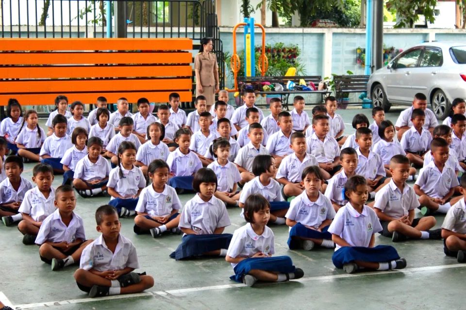 Des dizaines d'écoliers en uniforme blanc et bleu assis en tailleur sur un sol couvert, une enseignante debout derrière eux.