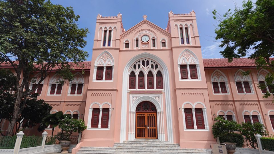 Pink neo-Gothic building façade with two crenellated towers, a clock, pointed-arch windows and a wooden door.