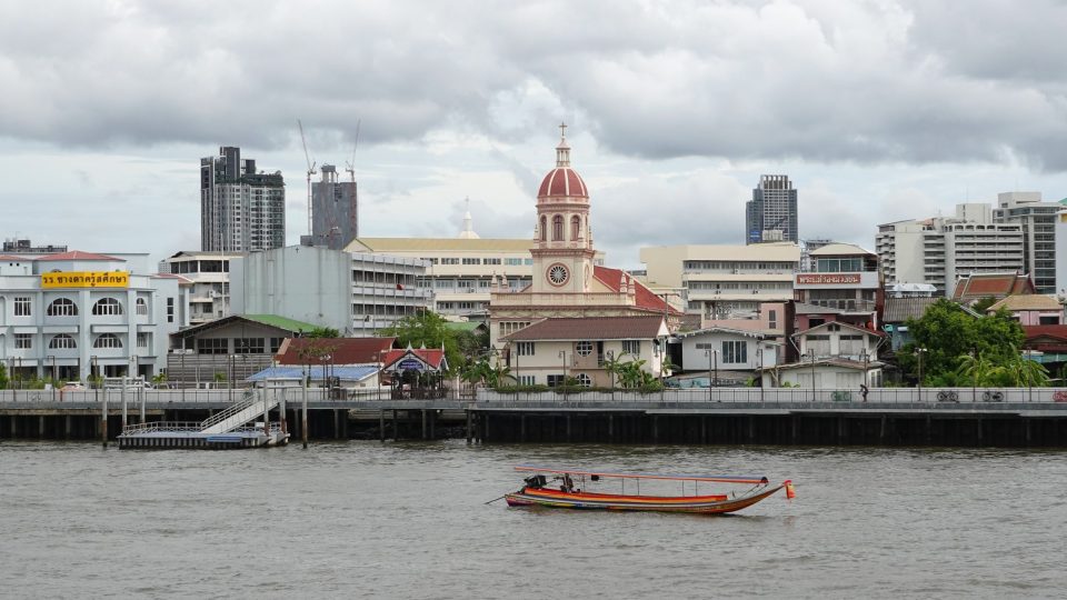 Church with a red dome and cream bell tower on a riverbank, longtail boat in front, tower blocks behind.