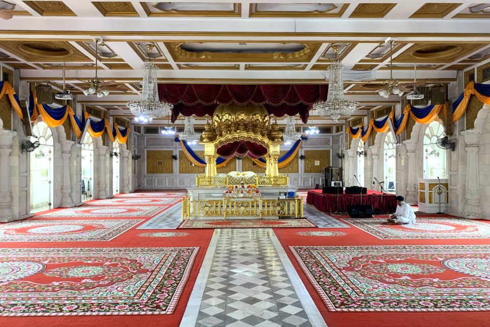 Prayer hall of a Sikh gurdwara in Pahurat: beneath the gilded canopy at the centre rests the Guru Granth Sahib, facing the carpets where worshippers sit on the floor. — © Wikimedia Commons Large Sikh prayer hall with red floral carpets, crystal chandeliers and a central gilded canopy sheltering the holy book.