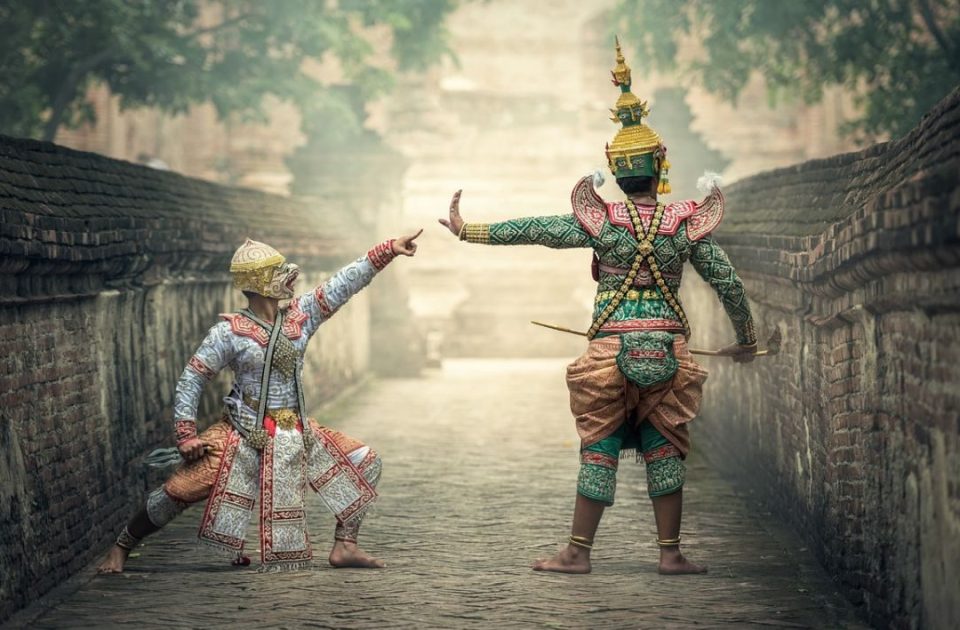 Two characters from khon, the masked dance-drama, in which every position of the hands, arms and legs carries a codified meaning. — © Mysakonnakhon.com Two dancers in khon costumes, one masked as a white monkey, one in green with a pointed crown, face each other on a brick-walled paved path.
