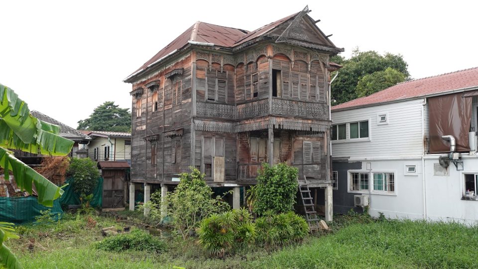 A timber house in Kudi Chin, raised on masonry pillars: note the pierced fretwork eaves and balustrades, louvred shutters and decorated gable, now in poor condition. — © Eric Monteil Old two-storey wooden house on pillars with fretwork friezes and a rusted metal roof, surrounded by vegetation.