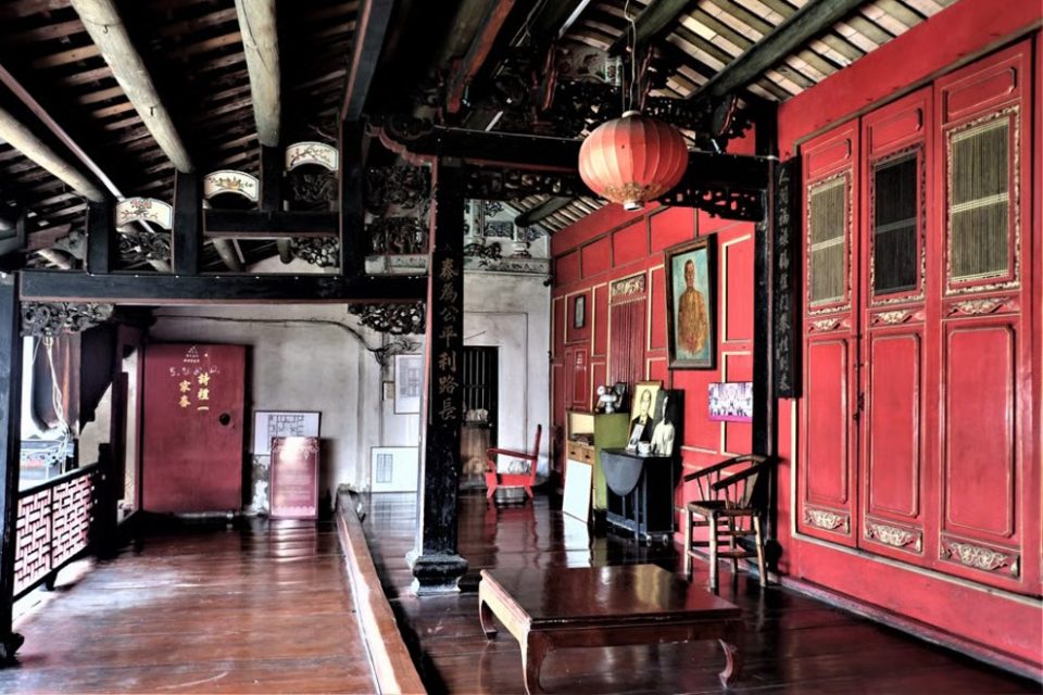 Interior of an old Chinese-style house with dark carved timber beams, red lacquered walls and doors, a red lantern and framed portrait.