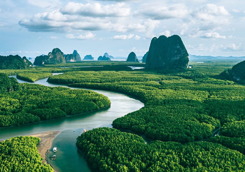 Aerial view of a winding river through dense mangrove forest, with limestone karst towers on the horizon and two small boats.