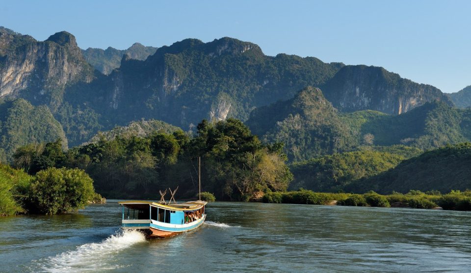 A blue and white wooden boat with a long roof travels along a wide river beneath forested karst mountains.