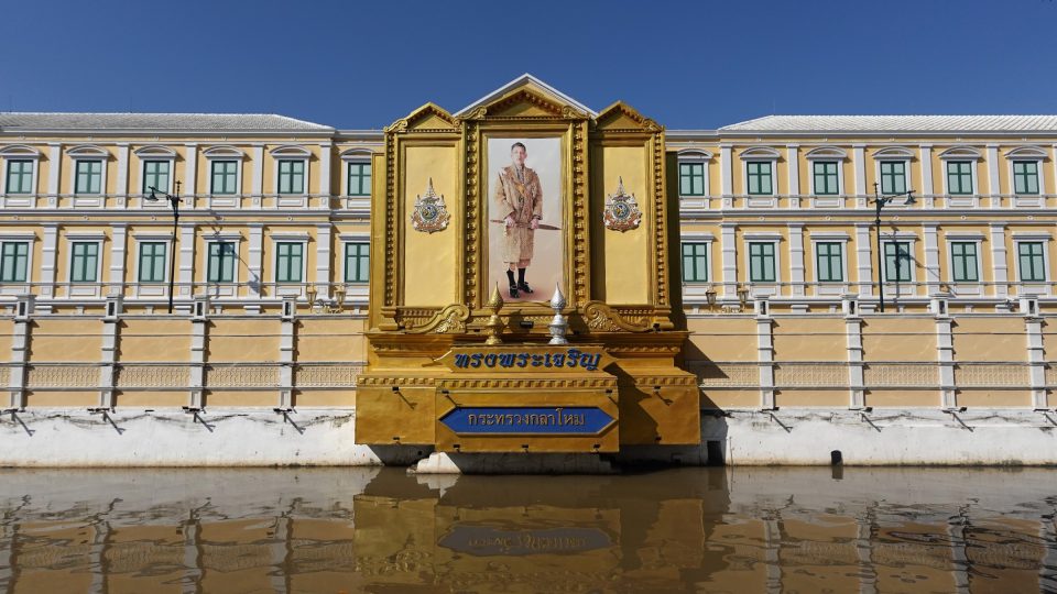 The Ministry of Defence on Rattanakosin Island: a gilded royal portrait stands at the edge of the khlong, before the yellow and green neoclassical facade. — © Eric Monteil Large royal portrait in a gilded frame before a long yellow facade with green shutters, reflected in canal water.