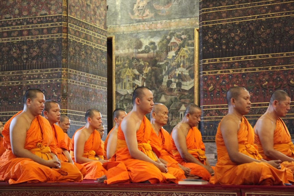 Monks in orange robes seated in a row, eyes half-closed, before floral-painted pillars and a mural.