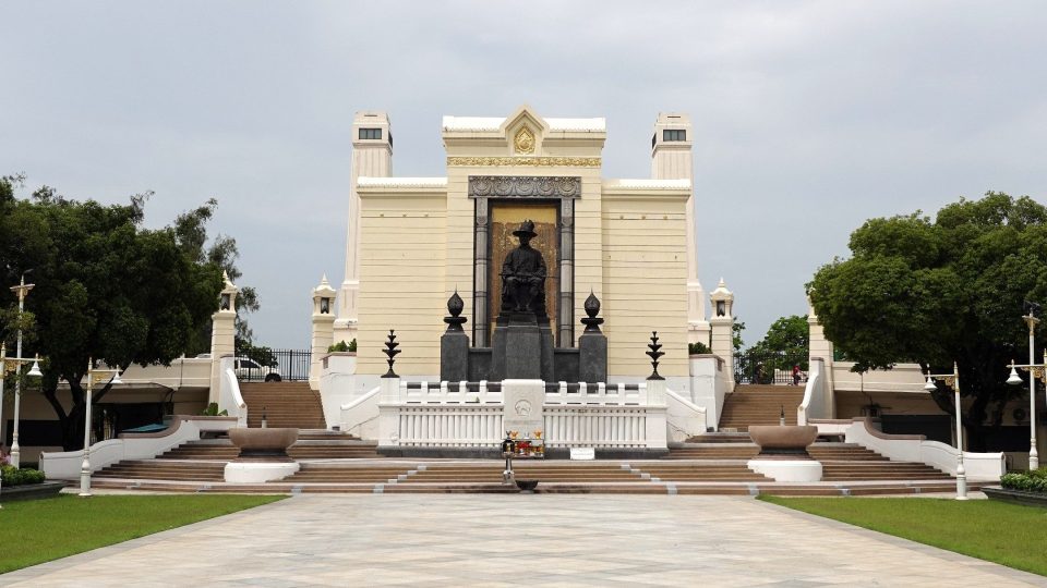 Bronze statue of a seated king before a cream portico with towers, fronted by steps, a white balustrade and a paved forecourt.