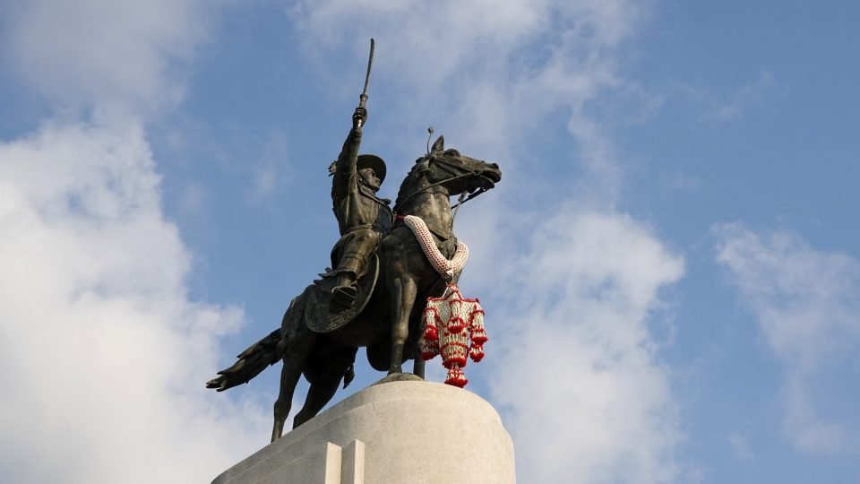 Bronze equestrian statue of king Taksin raising a sword, a flower garland around the horse's neck, on a pale stone plinth.