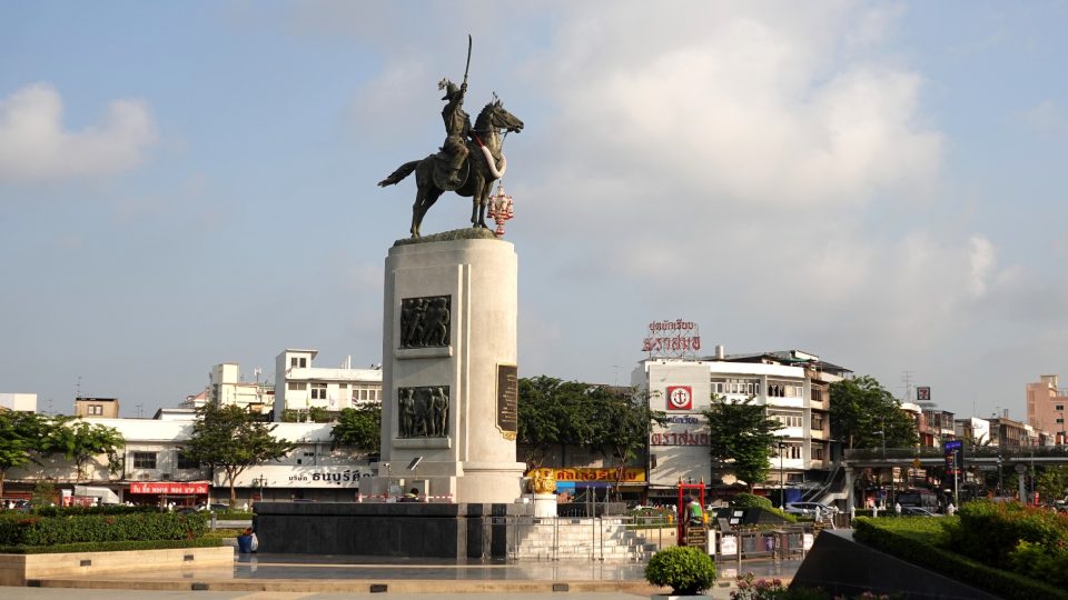 Statue équestre en bronze du roi Taksin brandissant une épée, sur un haut piédestal orné de bas-reliefs, au centre d'un rond-point urbain.