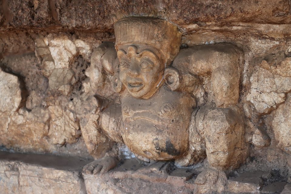 Carved squatting figure with raised arms, tall headdress and large ear ornaments, supporting a cornice above.
