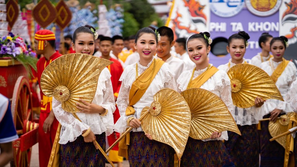 Dancers in traditional dress carrying golden fans in the Phi Ta Khon festival procession; a banner bearing the festival's name is visible behind them. — © Amazing Thailand Young women in white lace blouses and woven skirts holding large golden fans during a parade.
