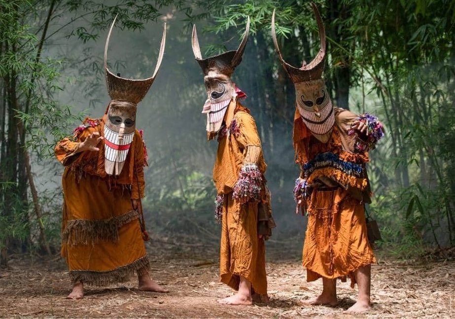Three Phi Ta Khon in costume: round-eyed masks with long teeth, topped by a basketry headdress with curved horns, and fringed outfits trimmed with coloured pompoms. — © Odasie Three masked figures with long-horned headdresses and fringed orange costumes pose barefoot in a bamboo grove.