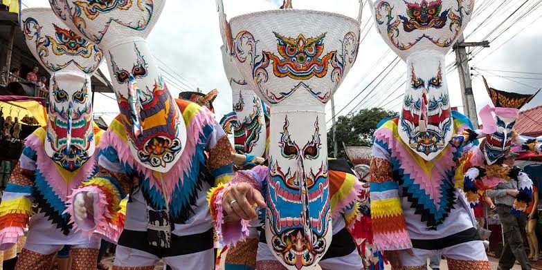 Costumed participants wearing large painted masks with long noses and tall headdresses, in multicoloured fringed clothing, on a street.