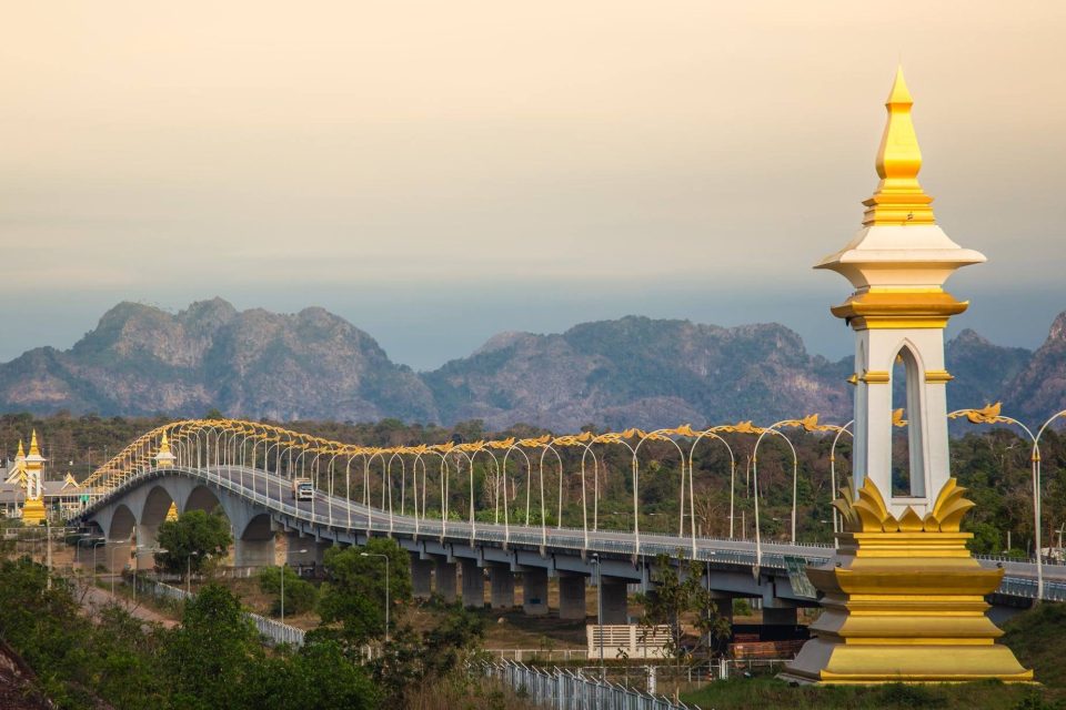 A road bridge spans the Mekong valley; its undulating golden lampposts and white-and-gold ornamental columns mark a border crossing, set against the limestone hills of the Lao bank. — © Jinnyz.com Long road bridge with golden arched lampposts, a white and gold column in the foreground and karst mountains behind.