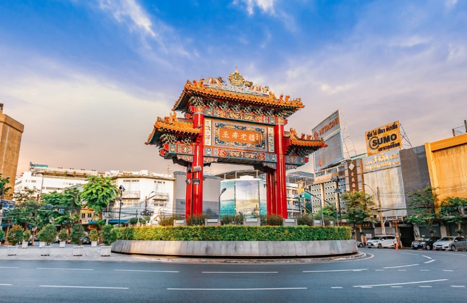 Large red Chinese gate with golden tiered roofs on a hedged roundabout, surrounded by buildings and shop signs.