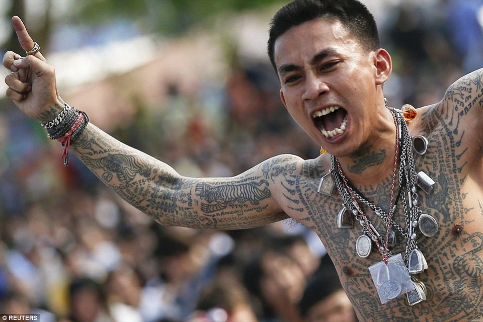 Bare-chested man covered in sacred tattoos, amulets round his neck, shouting with arm raised before a crowd.