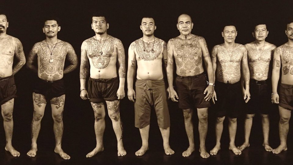 Men bearing sak yant across their chests, arms and legs: grids of script, protective figures and geometric patterns cover most of the body. — © Eric Monteil Eight bare-chested, barefoot men standing in a row against a black background, their torsos, arms and legs heavily tattooed.