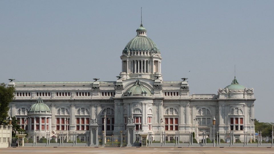 Large white marble neo-Renaissance building topped by a green central dome, fronted by railings and a broad paved square.