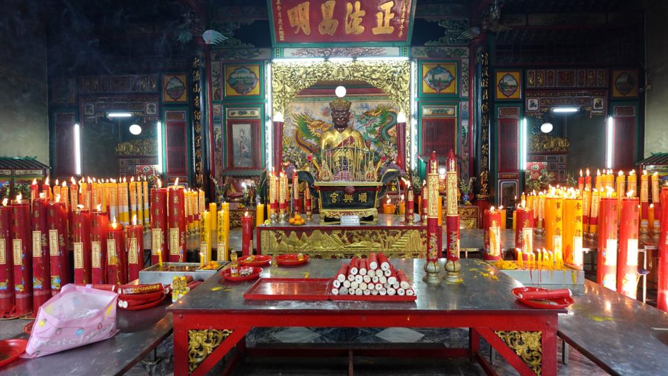 Interior of a Chinese shrine: gilded statue of a deity on an altar, surrounded by large lit red and yellow candles.