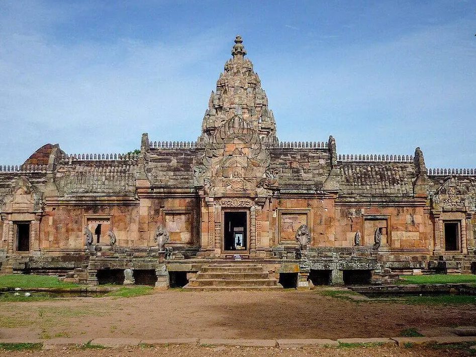 Khmer sandstone sanctuary: the enclosure gallery, pierced by an axial entrance pavilion, frames the carved central prang rising behind it. — © Buriram.live Pink sandstone Khmer sanctuary facade with a low gallery, central doorway and carved tower behind.