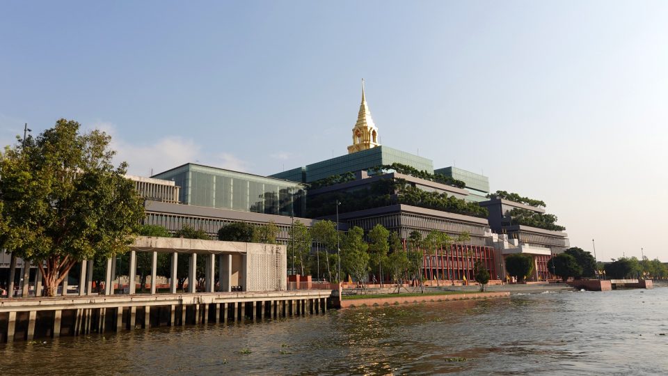 Large modern building with planted terraces topped by a golden spire, seen from a river, with a quay and trees in front.