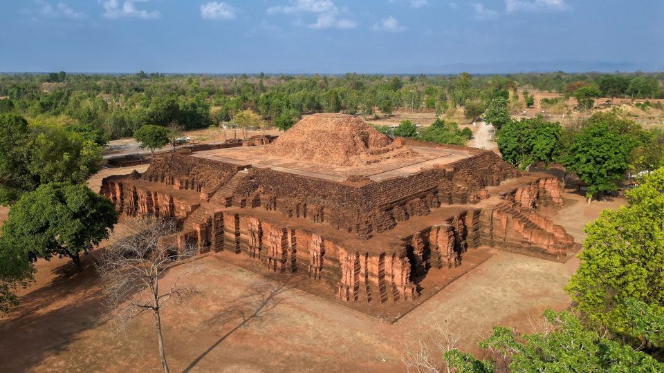 Large Dvaravati-period Buddhist monument in laterite: a square, indented base with axial stairways supports the levelled remains of a chedi. — © Salaween.blog Aerial view of a large square laterite monument with stepped terraces, topped by a brick mound, surrounded by trees.