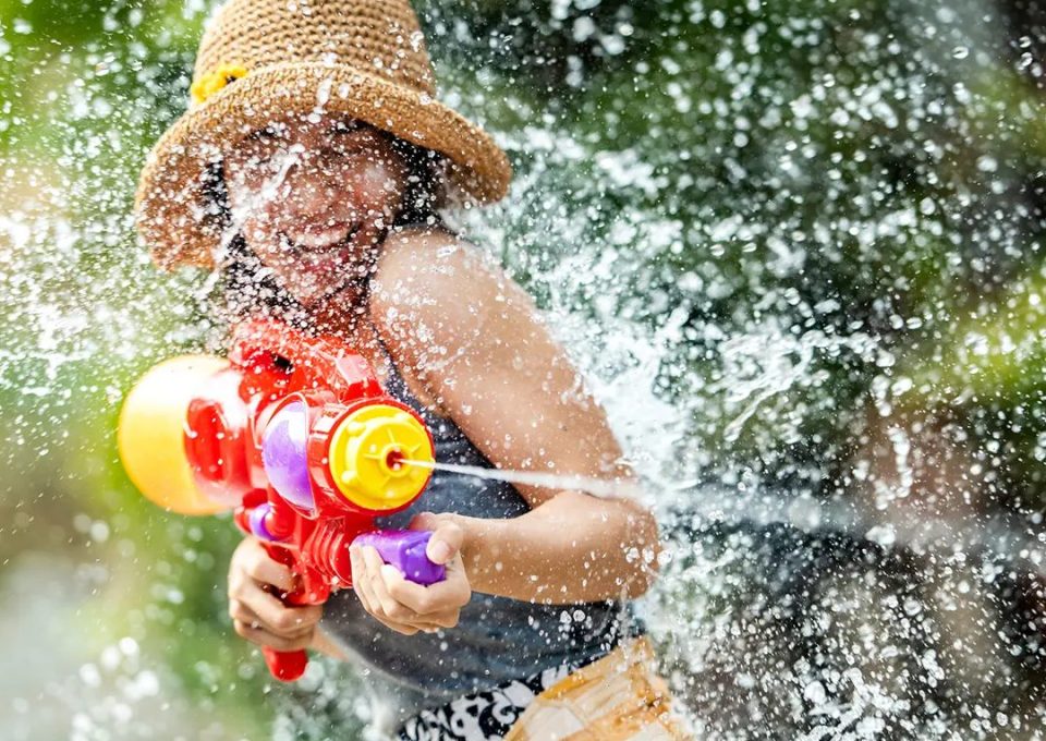 A woman in a straw hat fires a colourful plastic water pistol, surrounded by flying water droplets.