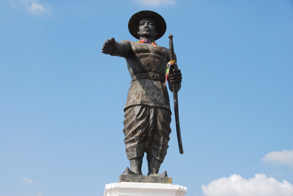 Statue of King Anouvong of Vientiane, arm outstretched and sword in hand, hung with garlands and ribbons left as offerings; this Lao ruler rose against Siam in 1826–1828. — © Flickr Bronze statue of a man in a wide hat, right arm outstretched, holding a sword against a blue sky.