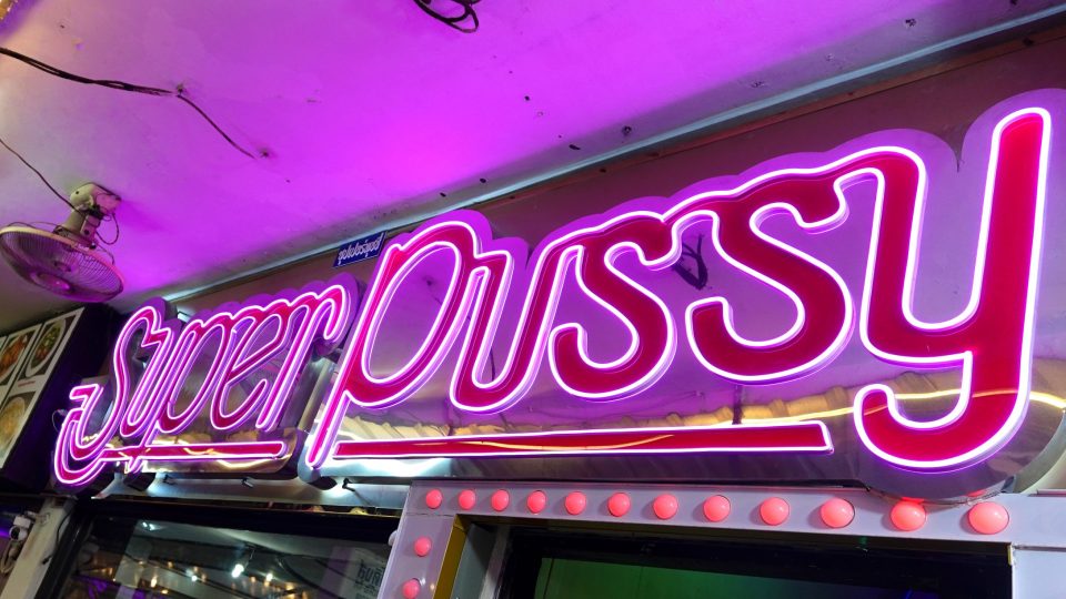 Pink and purple neon sign reading 'Super Pussy' above a bar entrance framed with red bulbs.