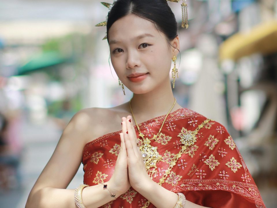 The wai: palms pressed together at chest height, fingers pointing upwards, accompanied by a slight bow and a smile; here worn with a traditional costume and a sash draped over one shoulder. — © Michelle Overseas Young woman in a red Thai costume embroidered with gold, wearing gold jewellery, smiling with palms pressed together.