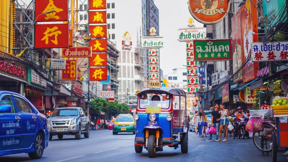Un tuk-tuk sur l'avenue Yaowarat, où les enseignes verticales en chinois et en thaï signalent commerces et bijouteries. — © TimeOut Un tuk-tuk bleu marqué « TAXI » roule dans une avenue bordée d'enseignes rouges à caractères chinois et thaïs.